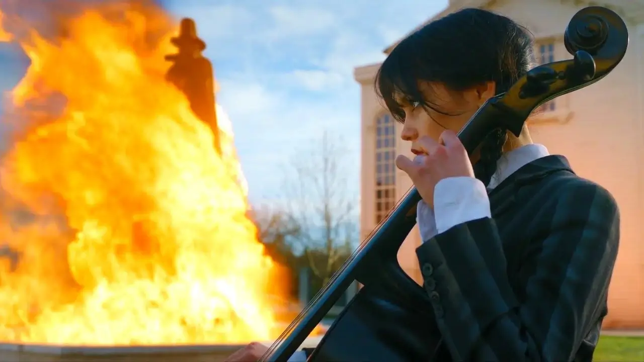 Mercredi (Wednesday) joue du violoncelle à côté d'une fontaine en flammes - scène extraite de la série